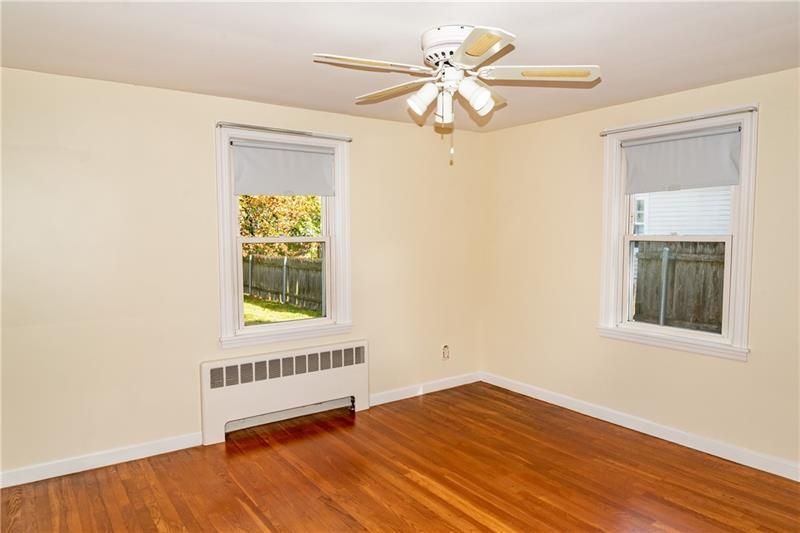 Empty bedroom with wood floors, two windows, radiator, and ceiling fan. Light yellow walls.