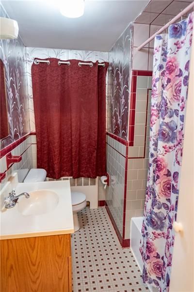 Small bathroom with white and red tile, wooden vanity, and a floral shower curtain.
