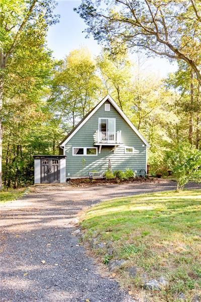 Green-sided A-frame house with small balcony on a wooded lot. Driveway and grass in foreground.