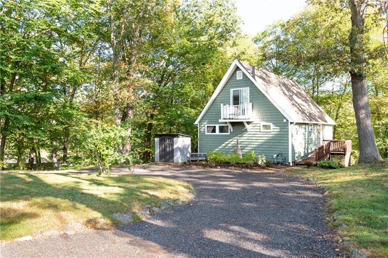 Green A-frame house with a small balcony, surrounded by trees and a gravel driveway.