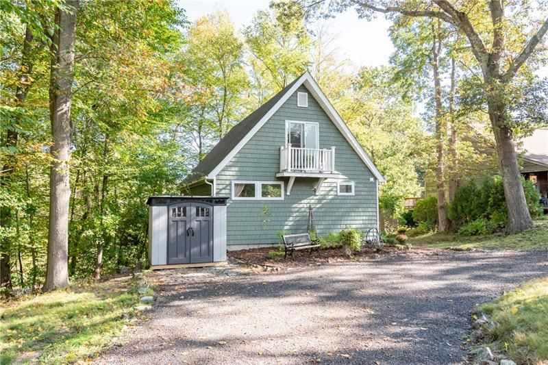 Green A-frame house with small balcony and storage shed, surrounded by trees. Gravel driveway.