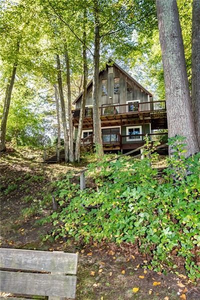 Wooden cabin nestled in trees on a hillside. Two-story structure with a deck. Green foliage and autumn leaves.