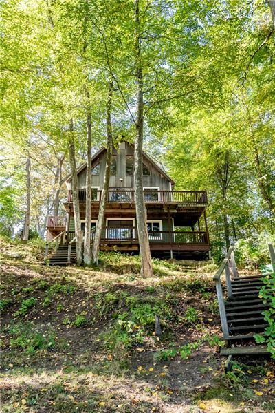 Two-story cabin nestled among trees on a hillside. Wooden deck, stairs to the lake.