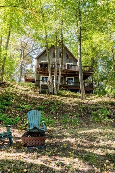 Cabin nestled in the woods with two-story deck. Blue Adirondack chair in front of fire pit.