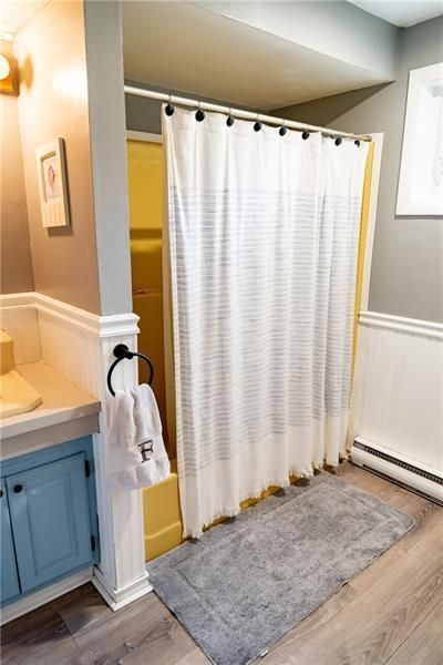 Bathroom with yellow shower, white curtain, gray rug, blue cabinet, and wood-look floor.