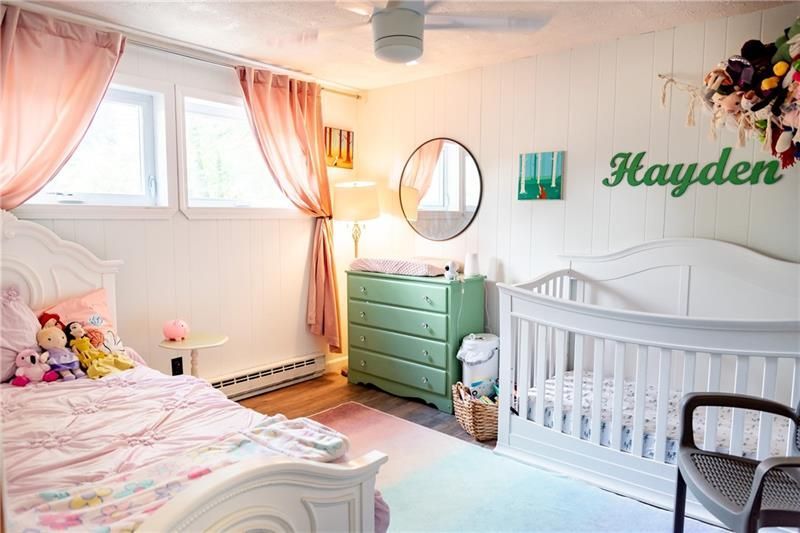 A nursery with a crib, bed, and changing table. Pink curtains and a green dresser add color to the room.