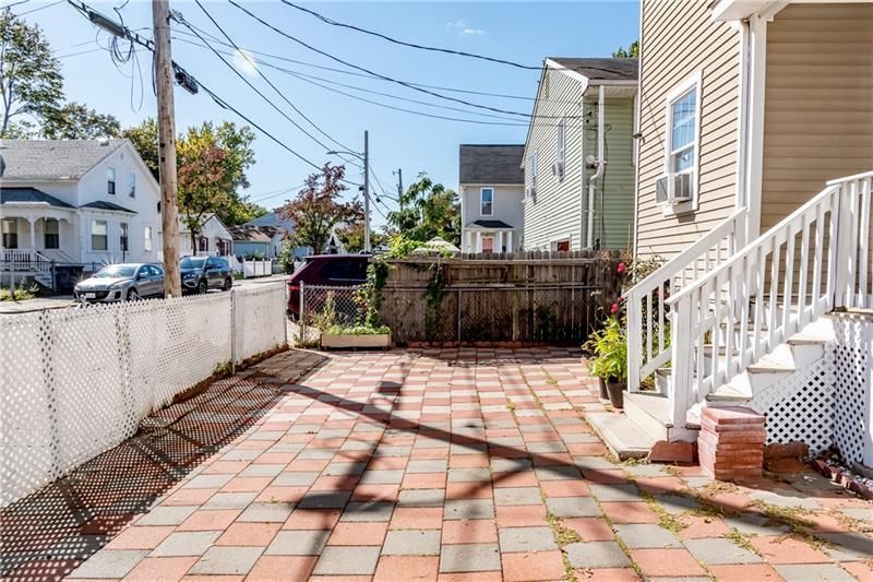 Brick patio and fenced backyard. Houses and car on a street. Sunny day.