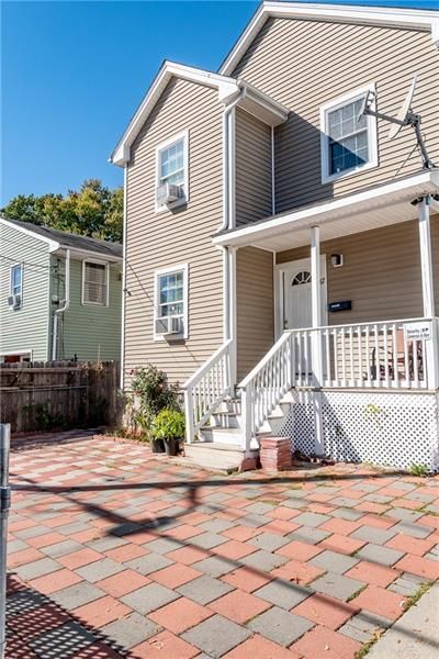 Beige two-story house with a porch and brick driveway on a sunny day.