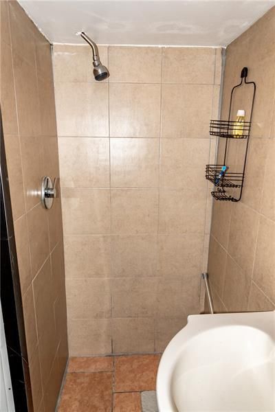 Small shower stall with tan tile walls, showerhead, and a white toilet. A black wire shelf holds toiletries.