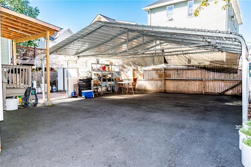 Large carport with metal roof over asphalt driveway. Wooden fence on the right, buildings in the background.