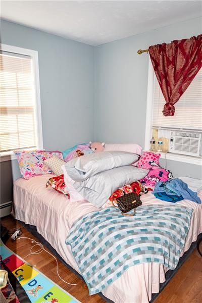 Bedroom with unmade bed, pink bed skirt, patterned linens, and a closed window with blinds.