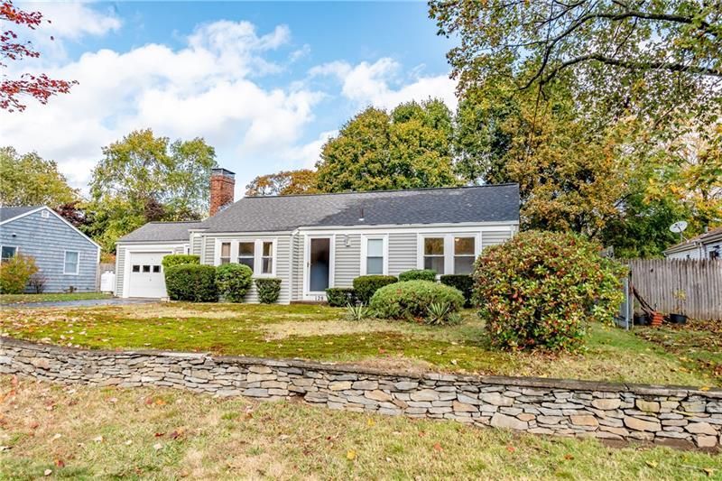 A gray single-story house with a small garage, bushes, and a stone wall in front.