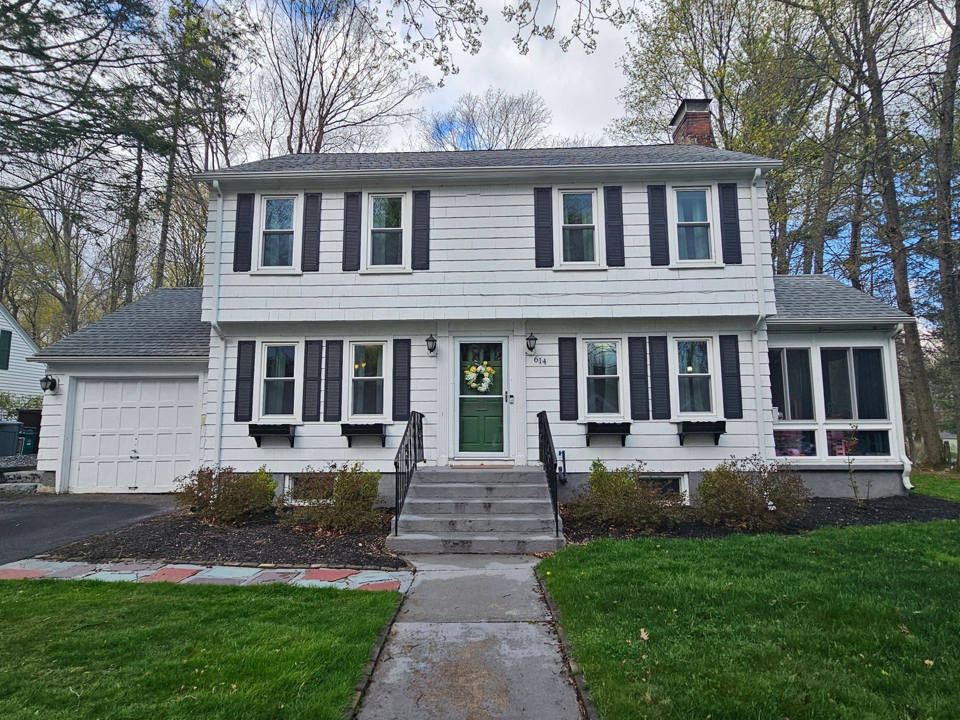 White two-story house with black shutters, green door, and gray roof. Front yard with green grass and a concrete path.