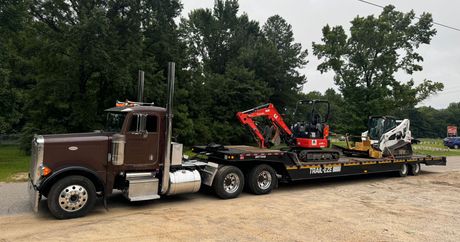 A semi-truck is carrying a small excavator on a flatbed trailer.