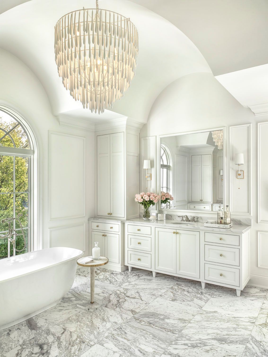 Bright white luxury bathroom with chandelier, vanity, freestanding tub, and large arched window