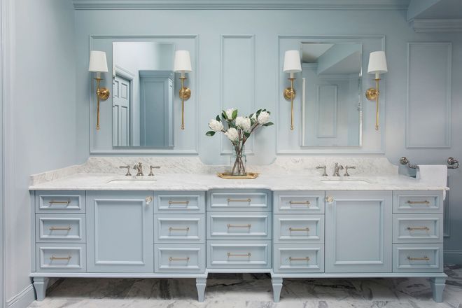 Elegant light blue bathroom vanity with double sinks, mirrors, sconces, and a vase of white flowers.