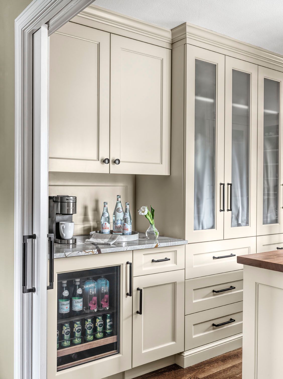 Built-in kitchen beverage station with cream cabinets, mini fridge, glass-front pantry, and countertop appliances.