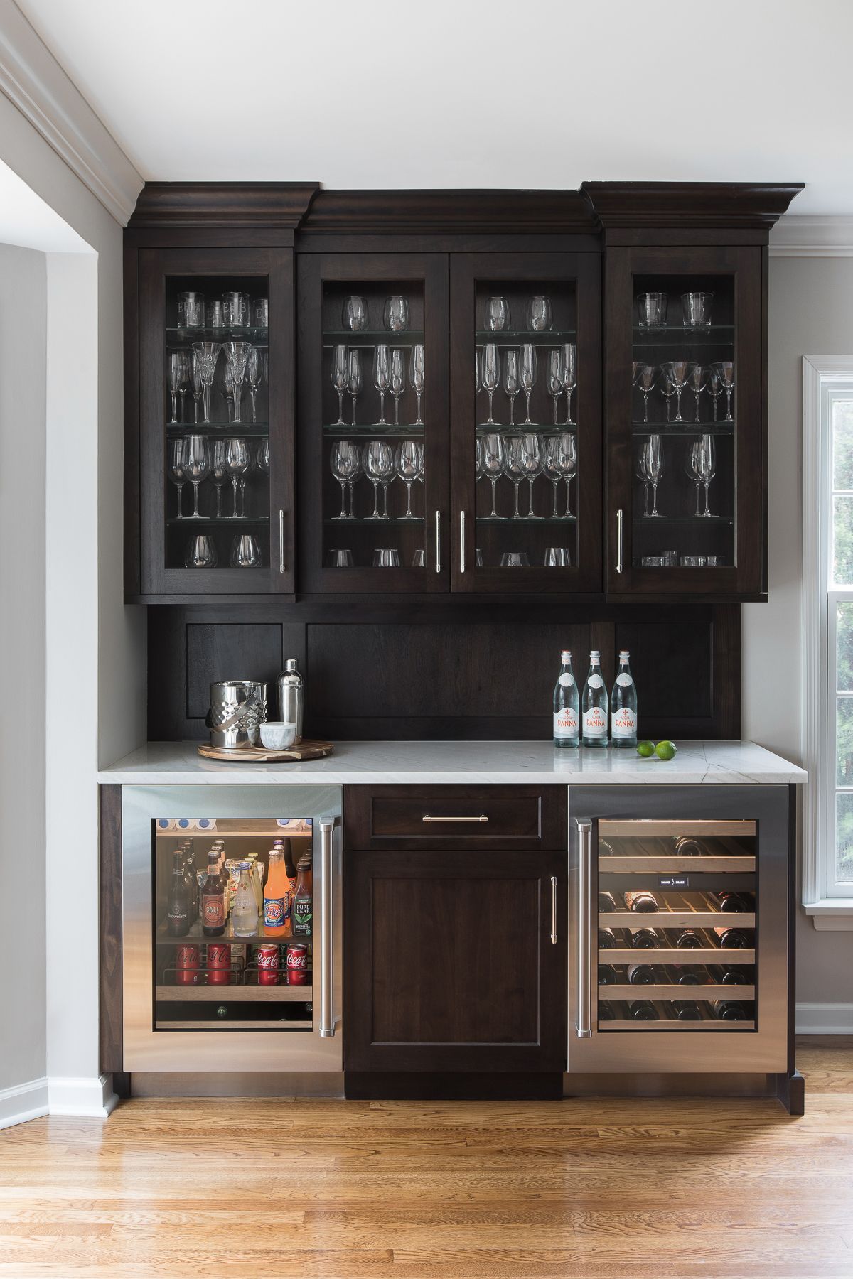 Dark wood home bar with glass cabinets, marble countertop, and wine cooler in a bright room.