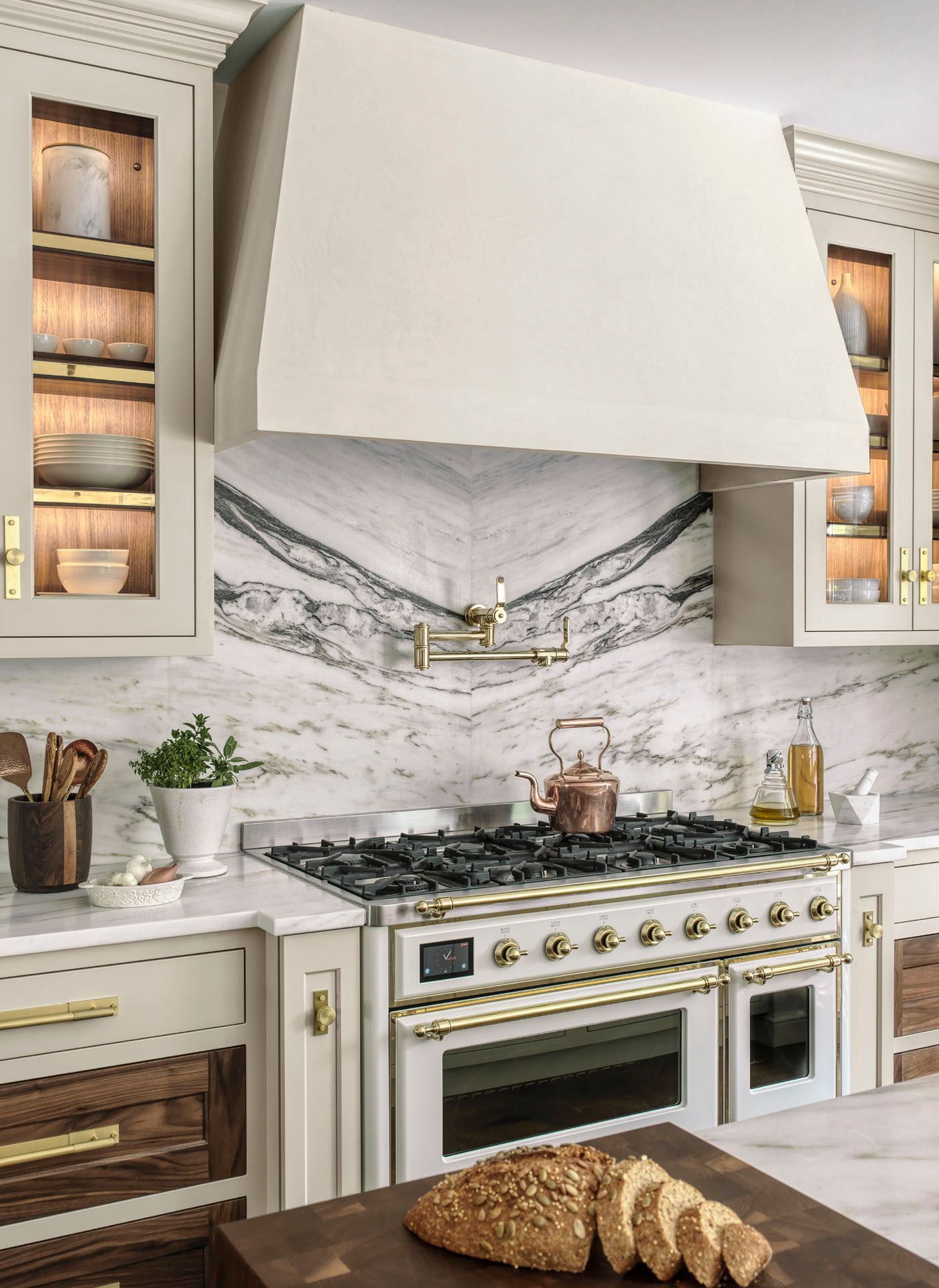 Elegant kitchen with marble backsplash, cream range hood, gold accents, and a loaf of bread on the counter.