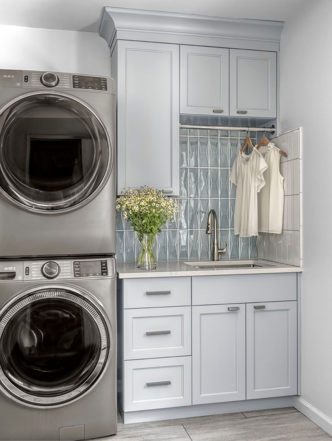 Laundry room with stacked stainless washer-dryer and white cabinets, countertop, and hanging clothes rack