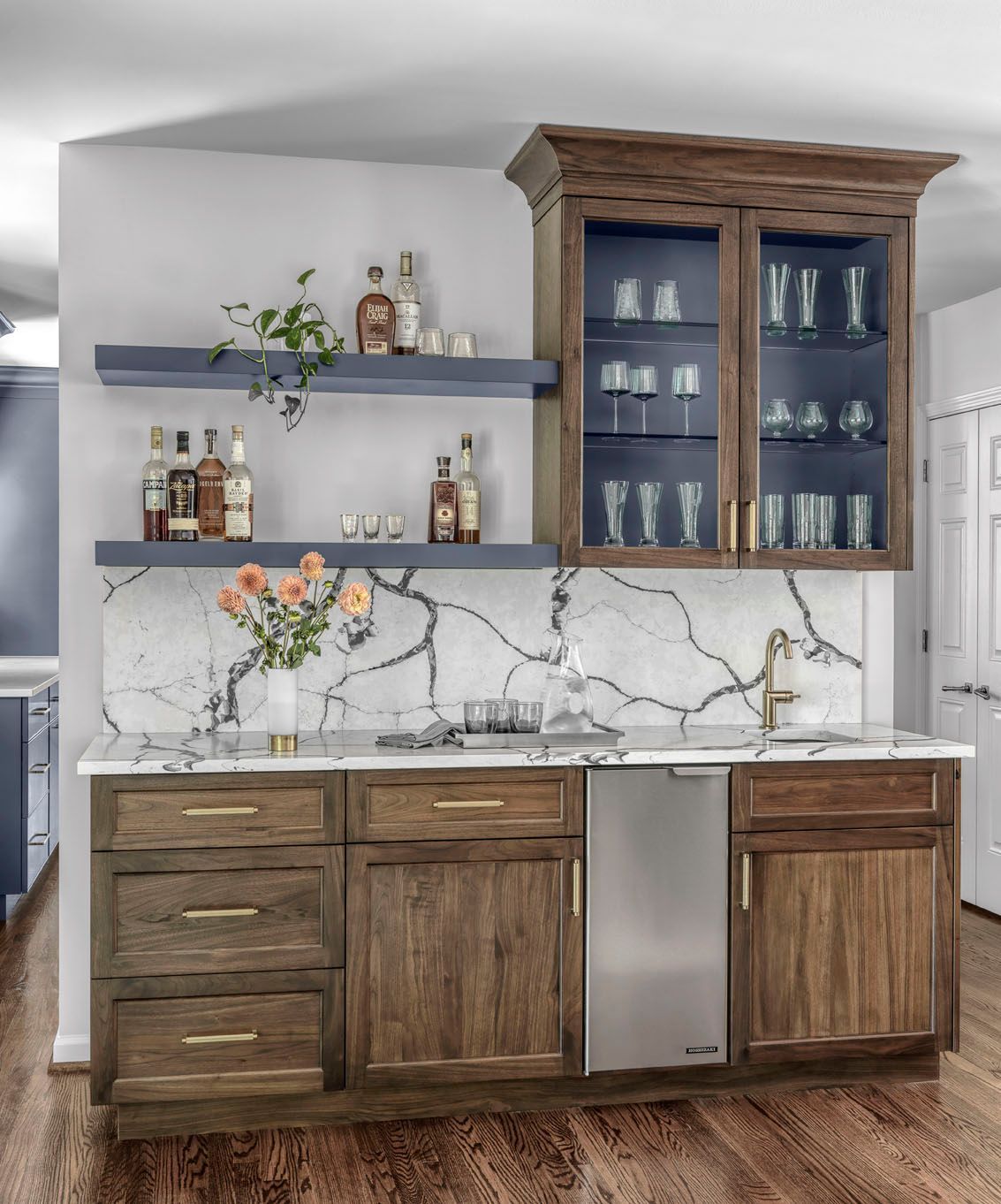 Modern kitchen bar with wood cabinets, blue shelves, marble backsplash, and stainless-steel refrigerator top.