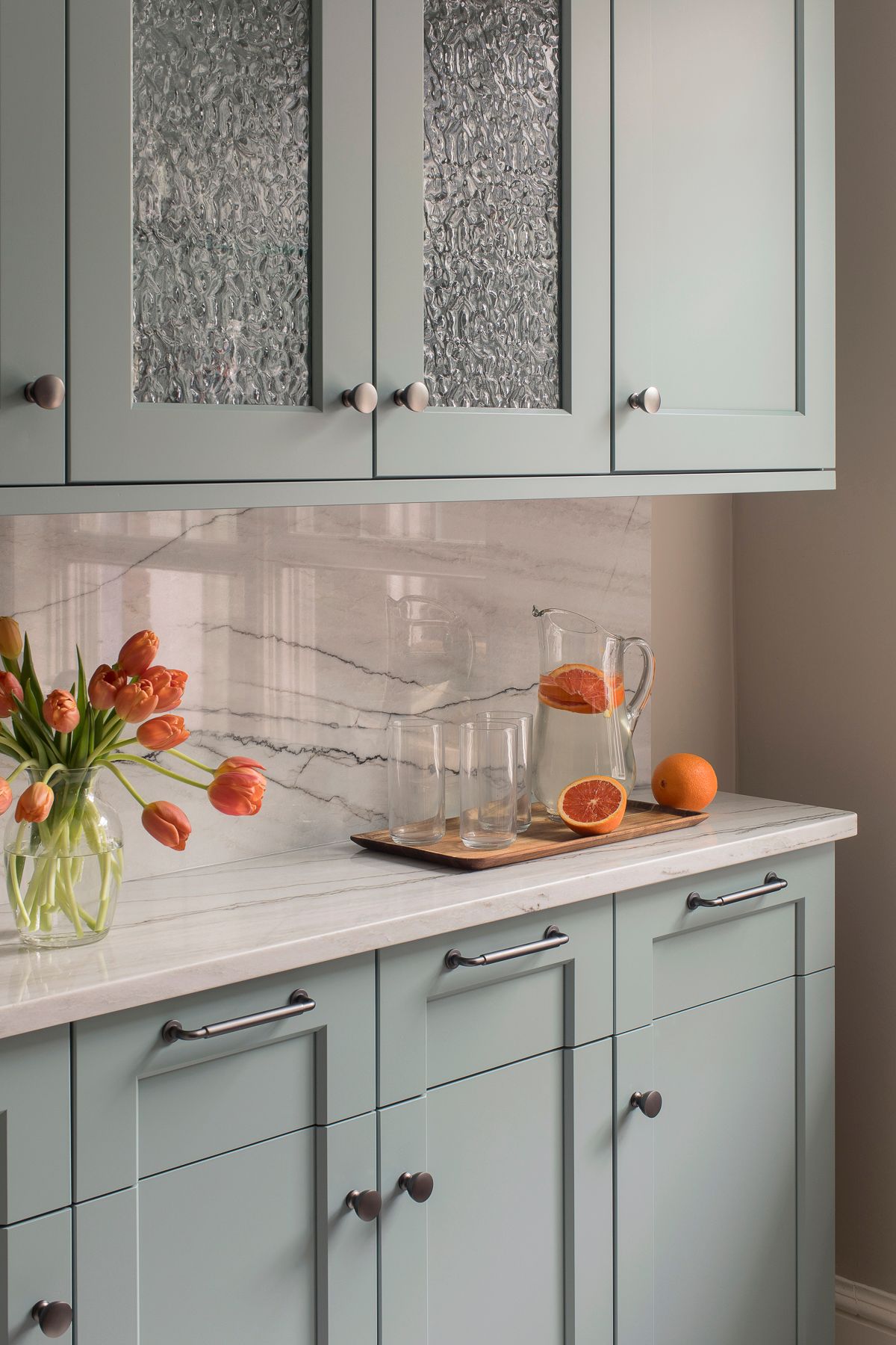 Pastel green kitchen cabinets with marble countertop, orange flowers, and a glass bowl of oranges