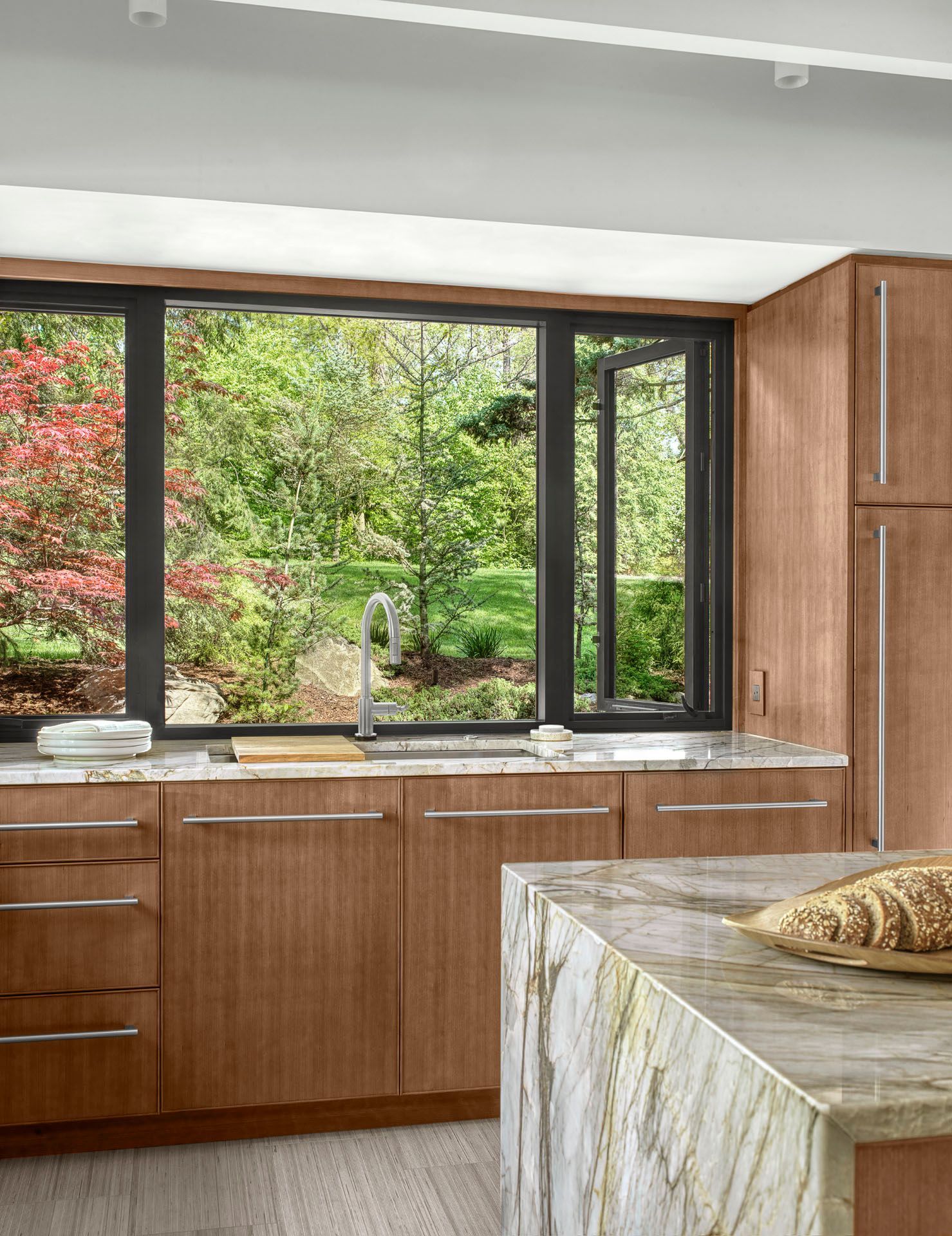 Modern kitchen with wood cabinets, large window, and marble island overlooking green trees outside