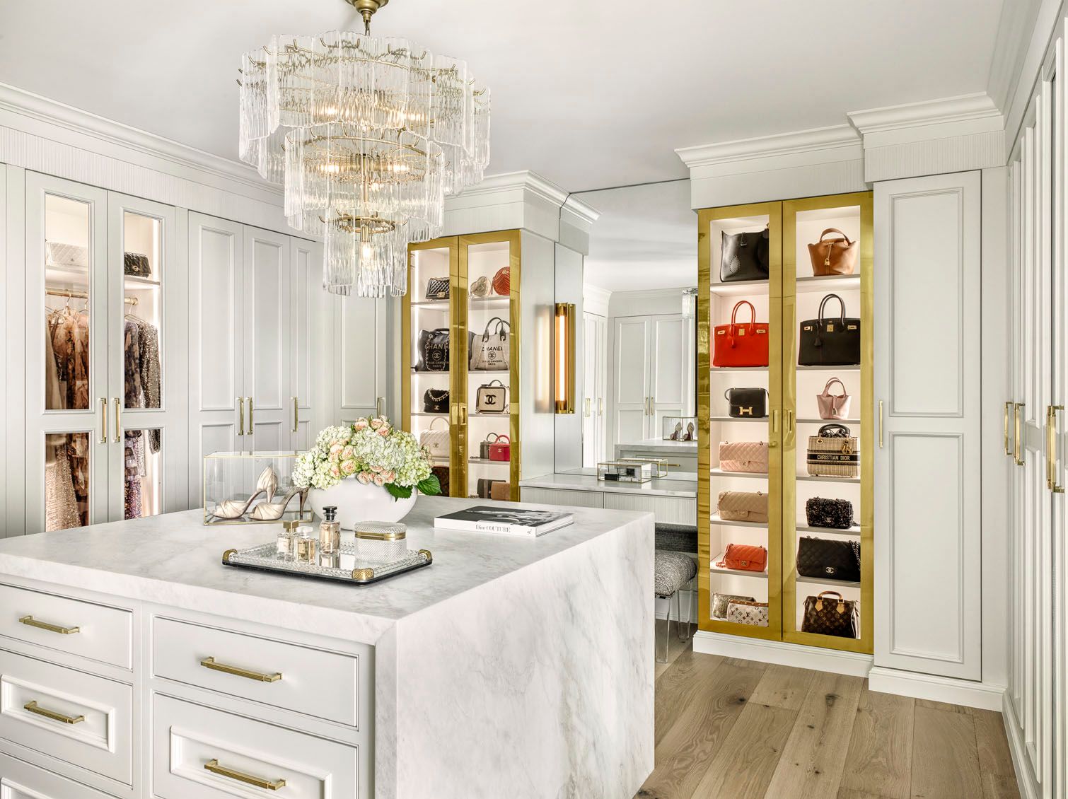 Elegant white kitchen with marble island, gold accents, glass-front cabinets, and crystal chandelier.