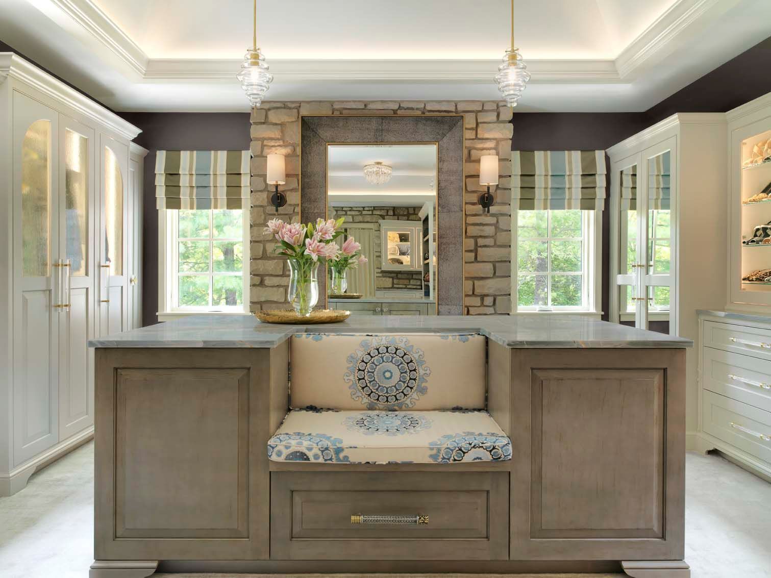Bright kitchen with central window seat, stone fireplace, white cabinetry, and floral vase between windows