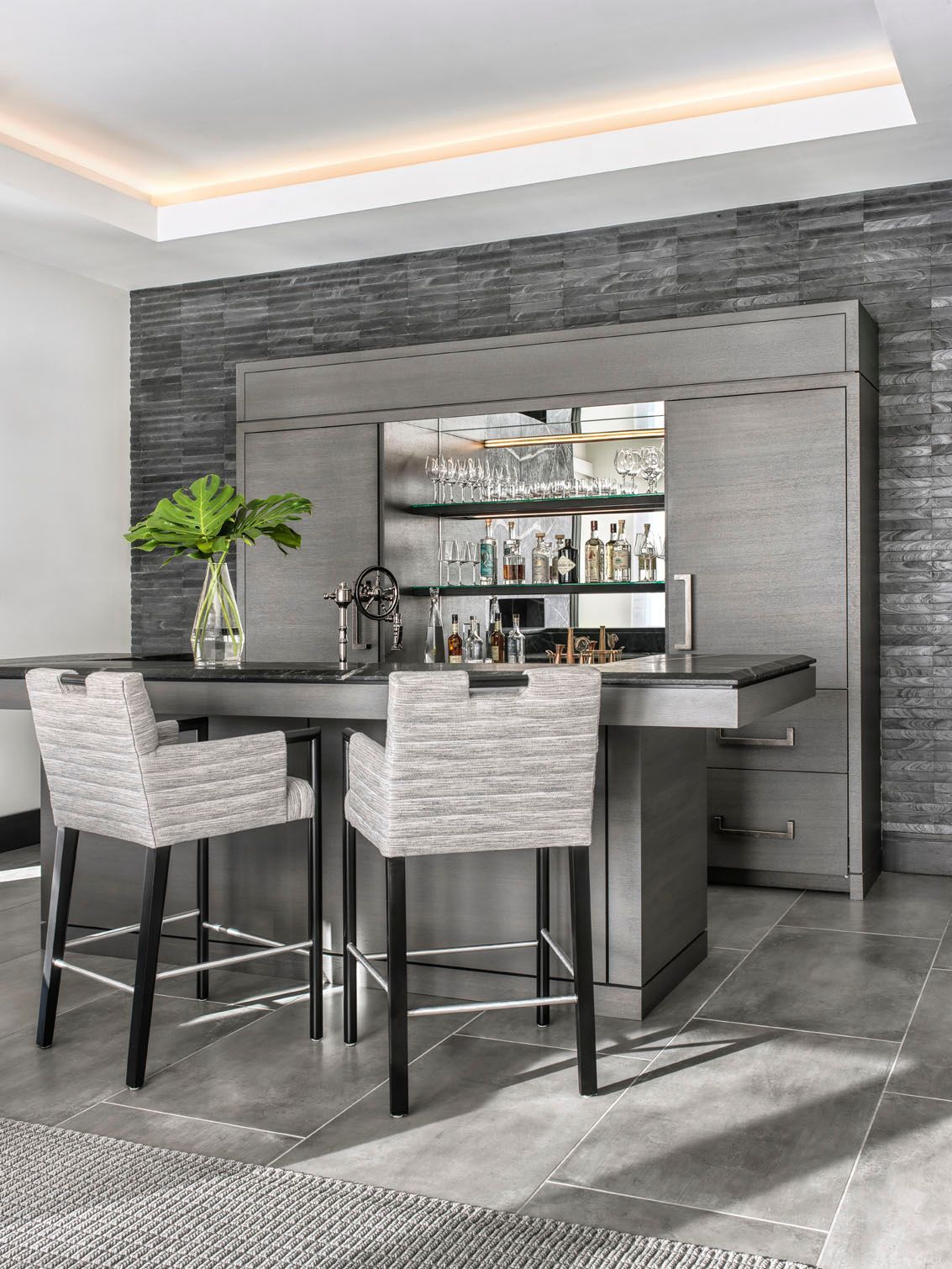 Modern gray kitchen with island, two upholstered stools, and a mirrored bar area with a plant