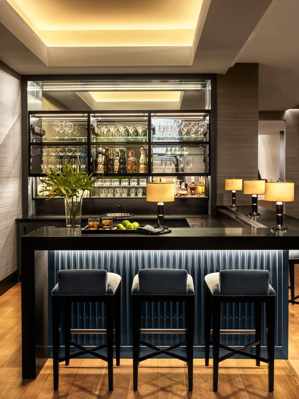 Modern hotel bar with black counter, four striped stools, warm lighting, and shelves of bottles