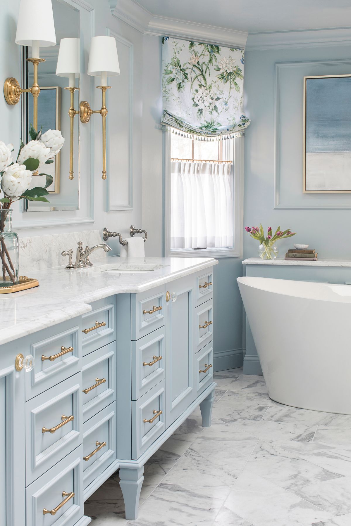 Light blue bathroom with marble floor, white vanity, brass sconces, and a freestanding tub.