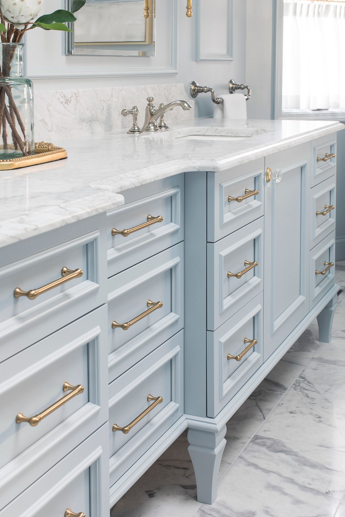 Light blue bathroom vanity with marble countertop, gold handles, and dual sinks beneath a mirror
