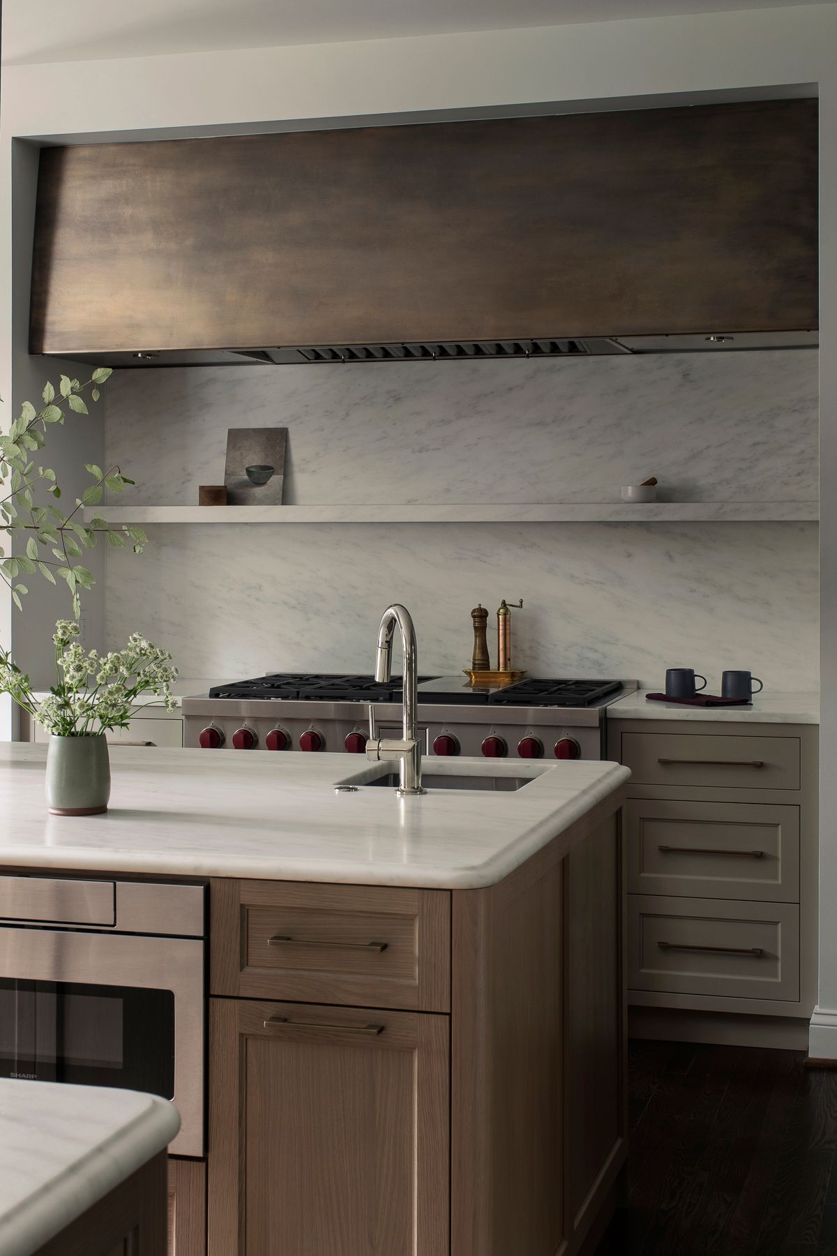 Modern kitchen with large hood, open shelves, white island, and sink faucet in front of a stove.