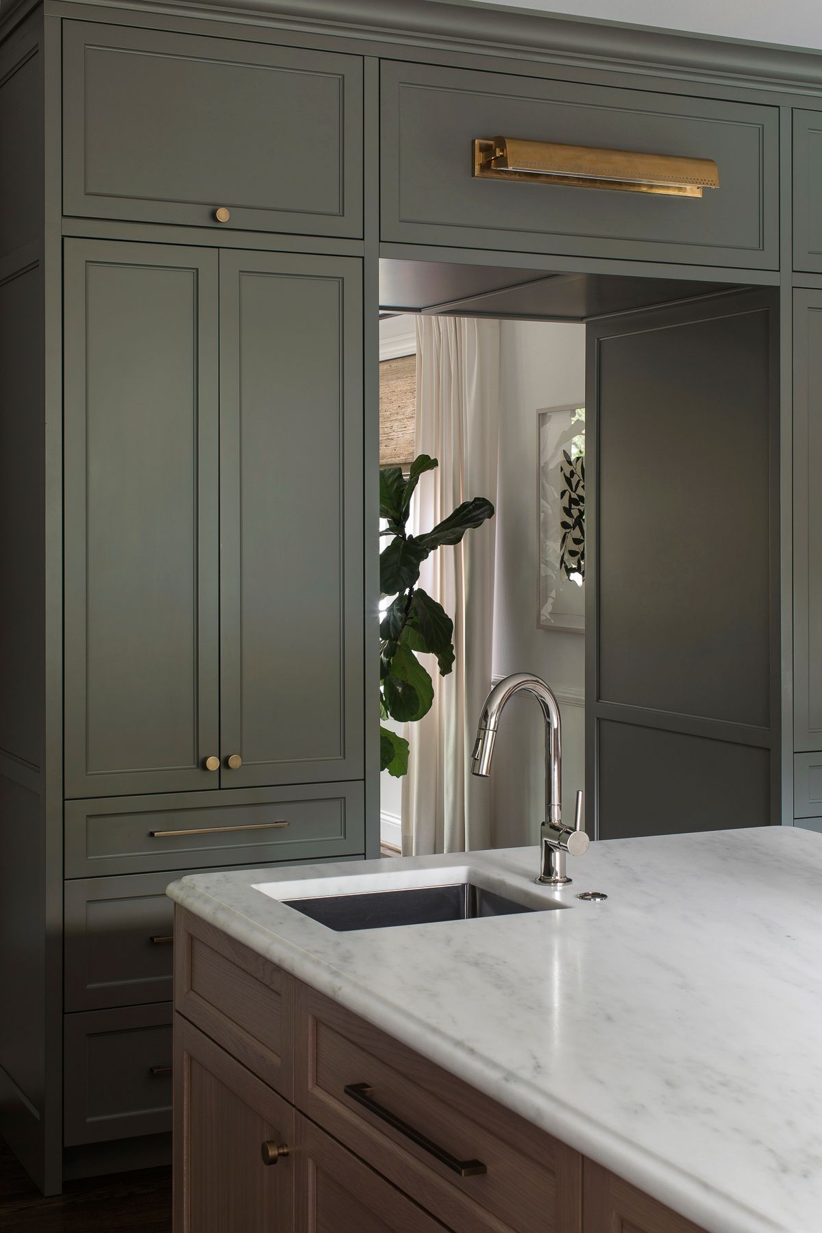 Modern kitchen with green cabinetry, marble island, and stainless faucet by a large doorway.