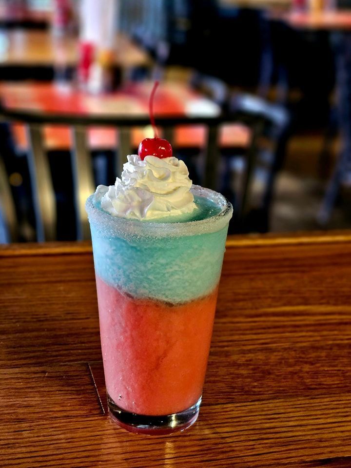 A red, white, and blue drink with whipped cream and a cherry on top is on a wooden table.