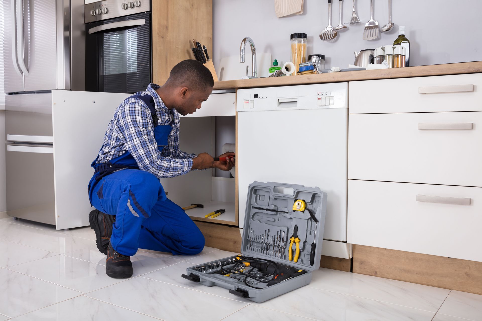 A service technician in blue work overalls kneels on a kitchen floor, repairing an appliance with a toolbox nearby.