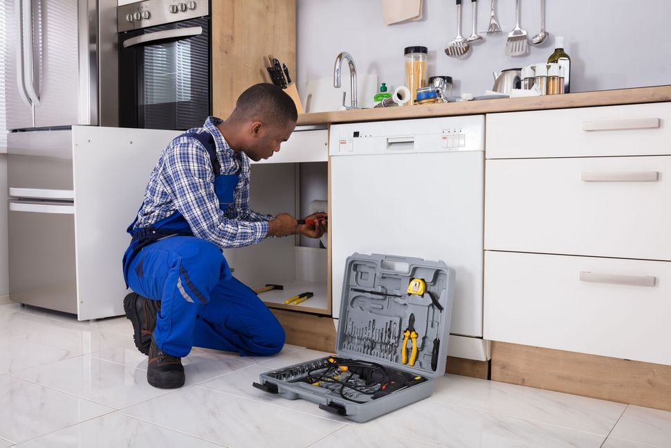 A service technician in blue work overalls kneels on a kitchen floor, repairing an appliance with a toolbox nearby.