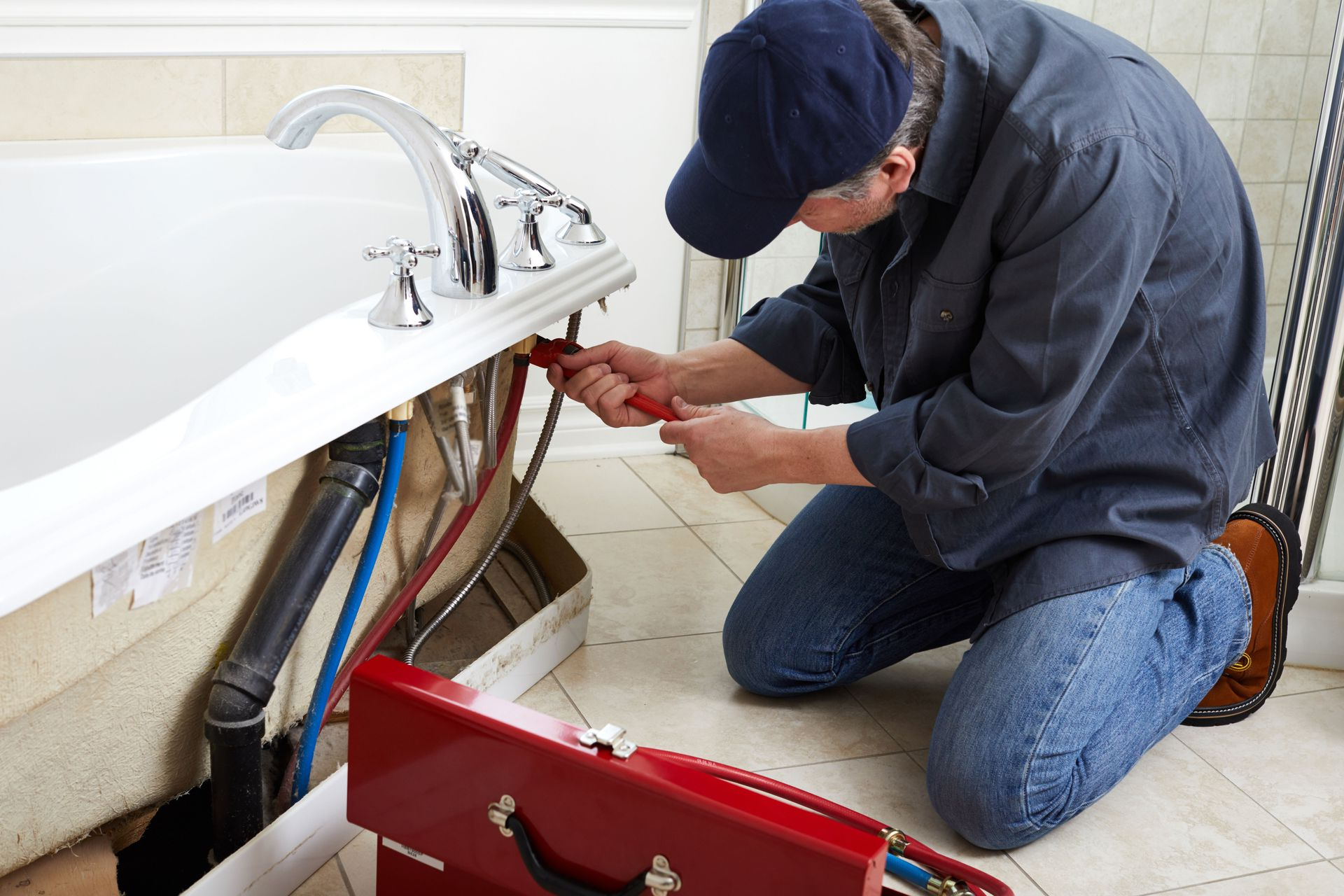 A plumber in a blue cap kneeling on a tiled floor to repair plumbing underneath a white bathtub.