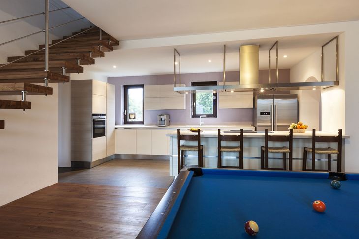 Modern kitchen with island and pool table in foreground. Wooden staircase on the left, stainless steel appliances, pale lilac walls.
