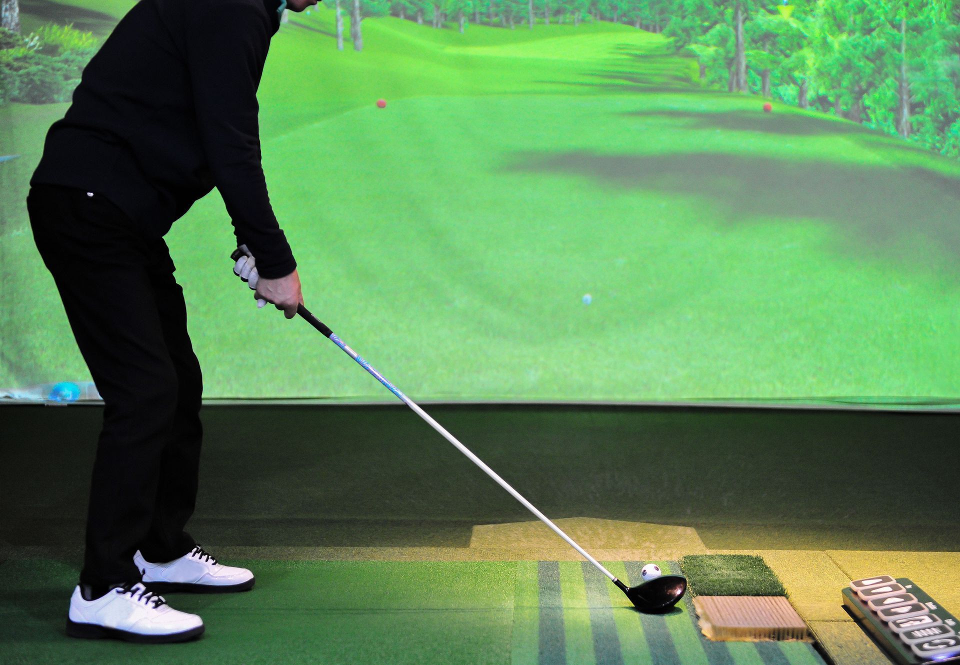 Person in black clothing swings a golf club at a tee in an indoor golf simulator.