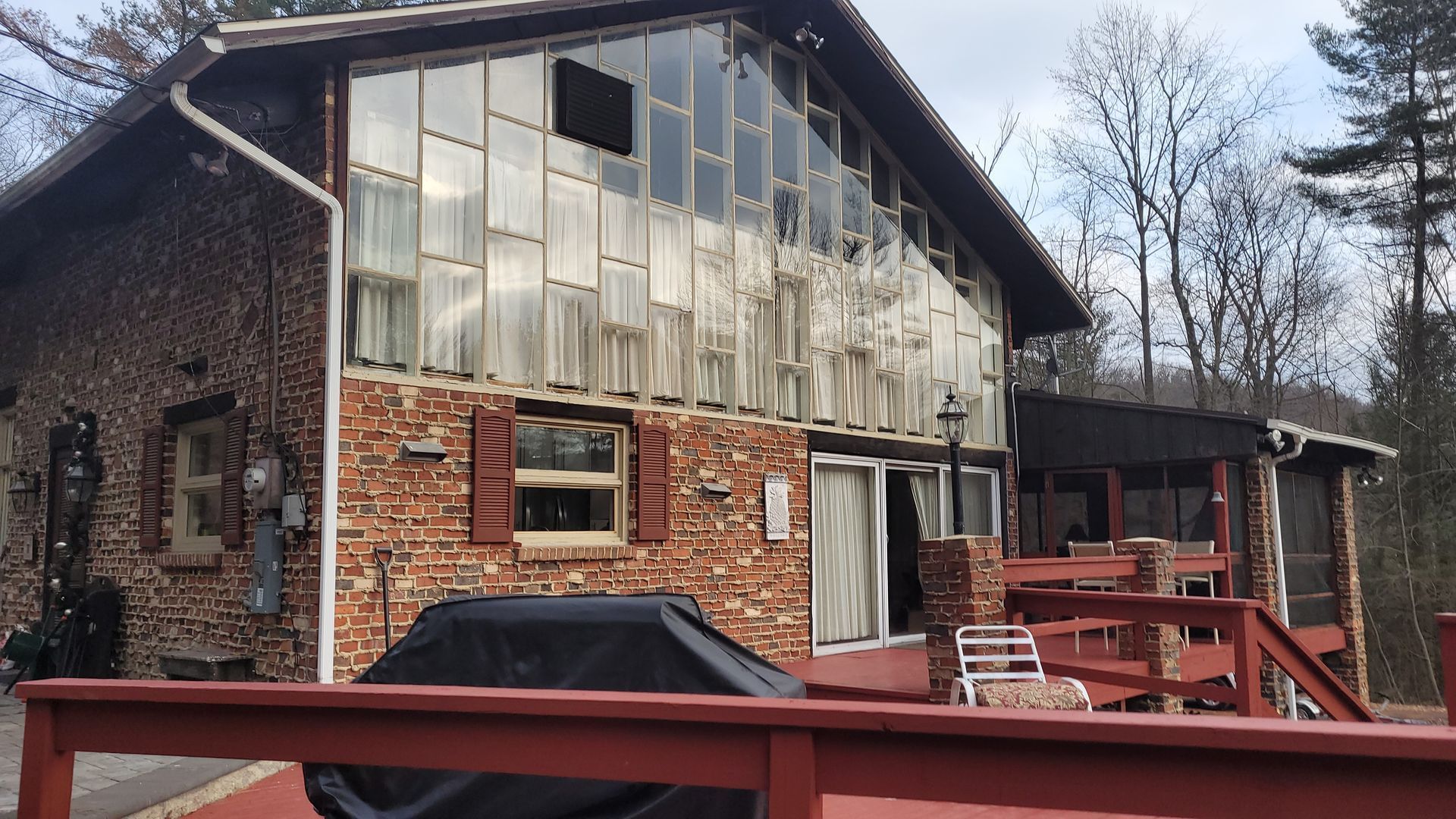 Brick house with large glass wall, red deck, and covered porch in a wooded setting.