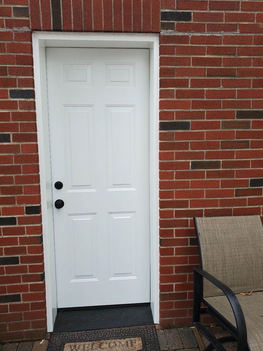 White door set into brick wall, black doorknobs, welcome mat, and patio chair.