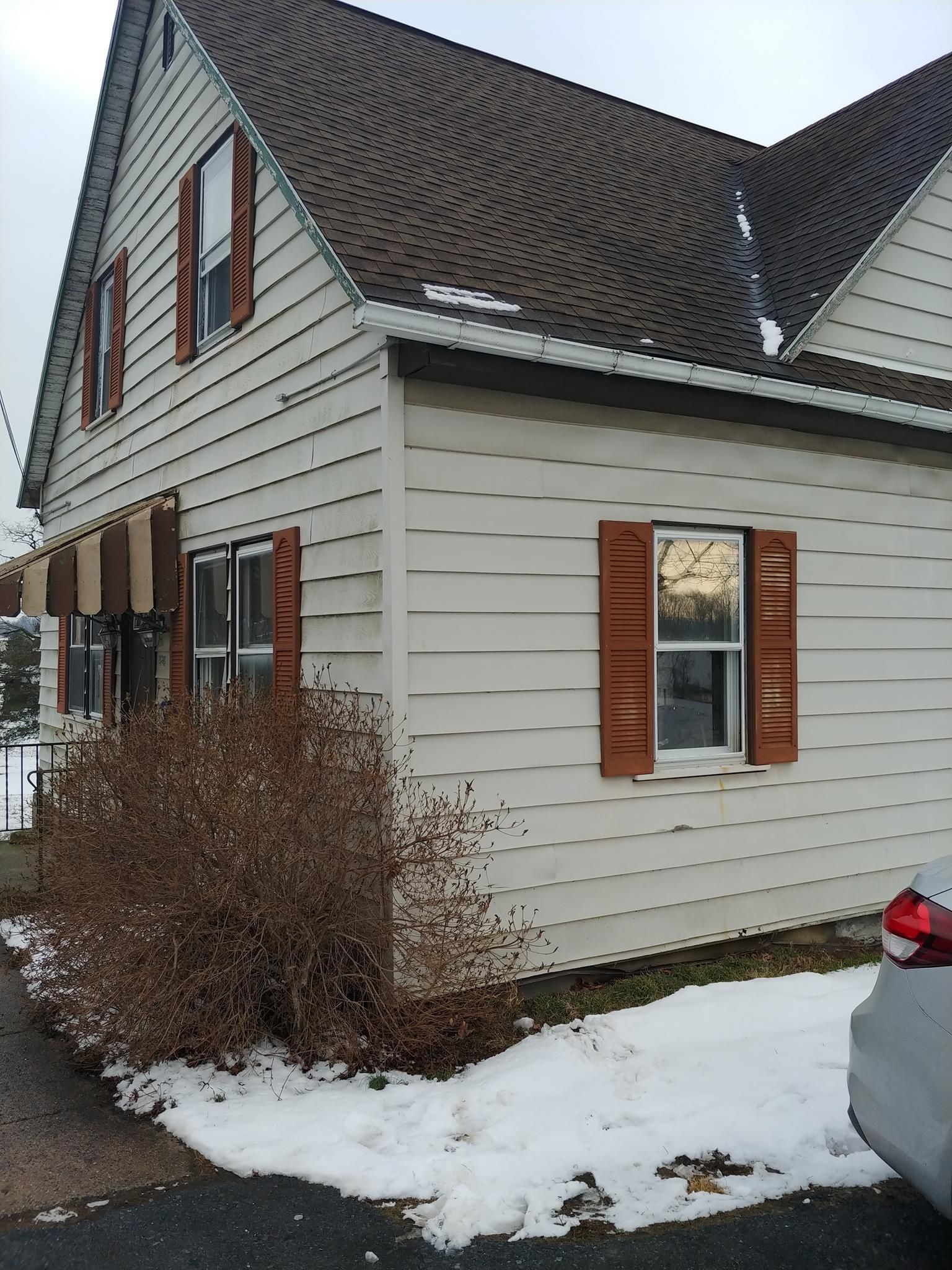 Beige house with brown roof and shutters, snow on the ground, overcast sky.