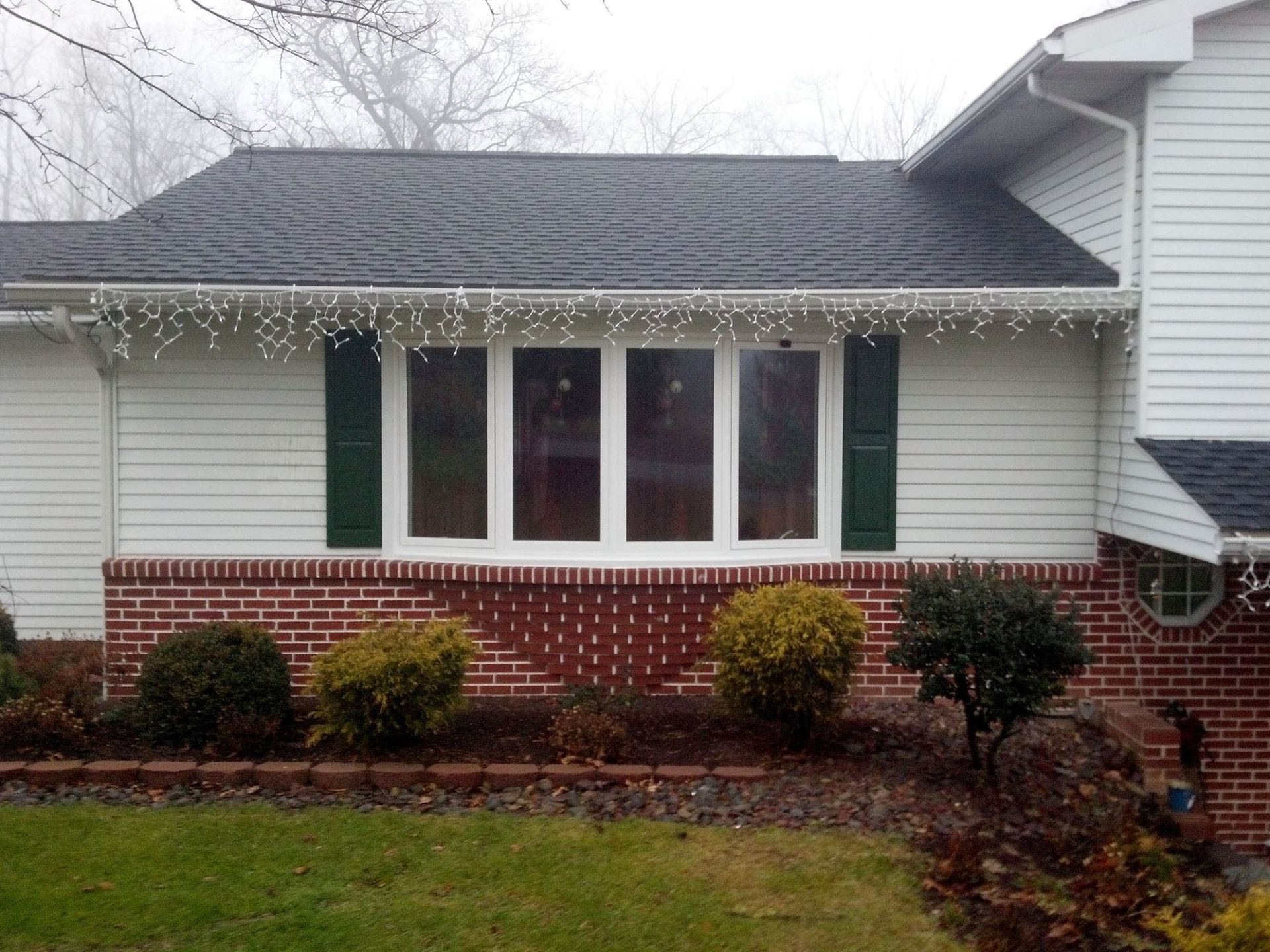 White house with black roof, large windows, green shutters, and brick base.  White siding, green lawn.