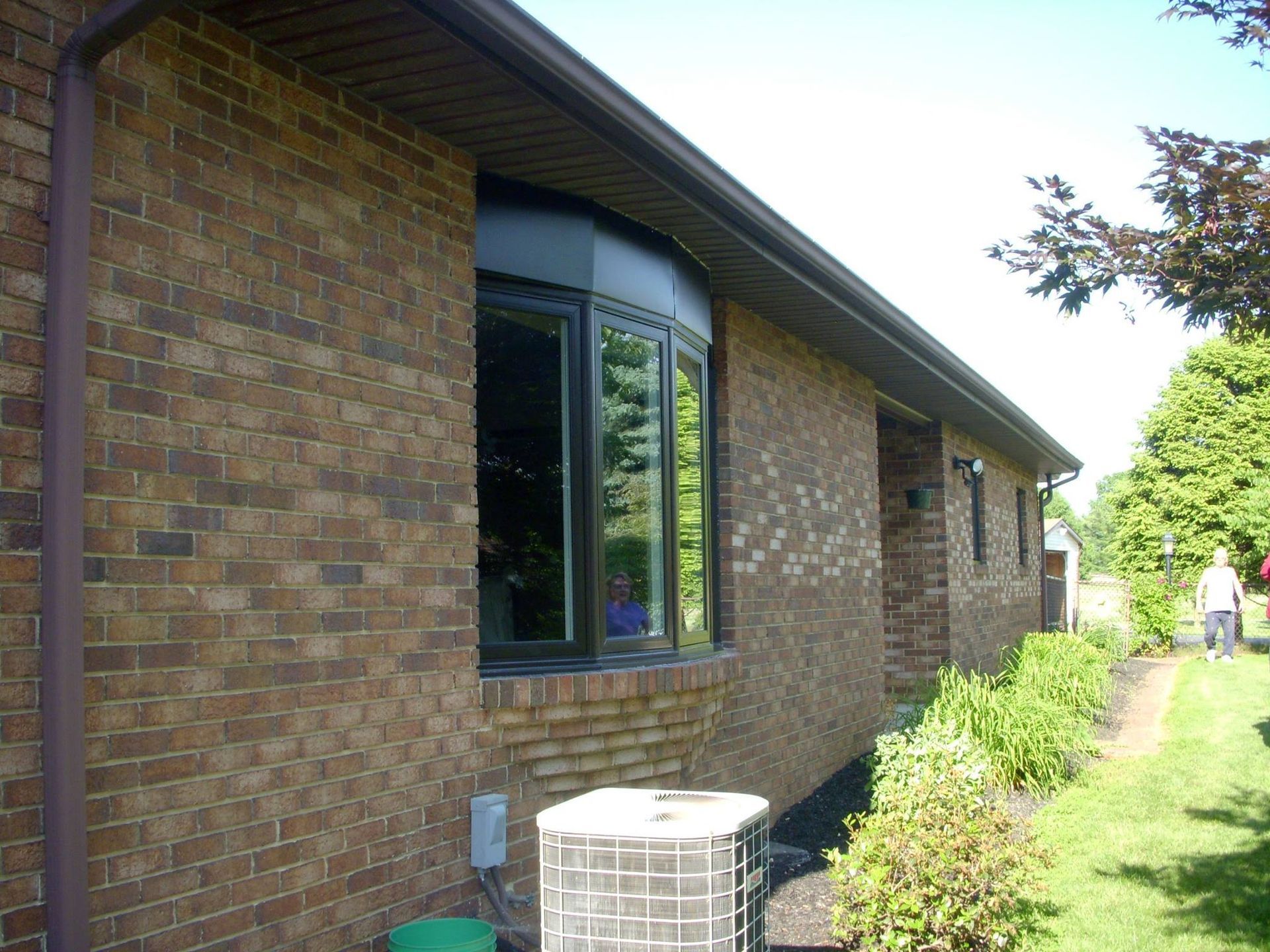 Brick house with a bay window, a/c unit, and greenery; person walking on lawn.