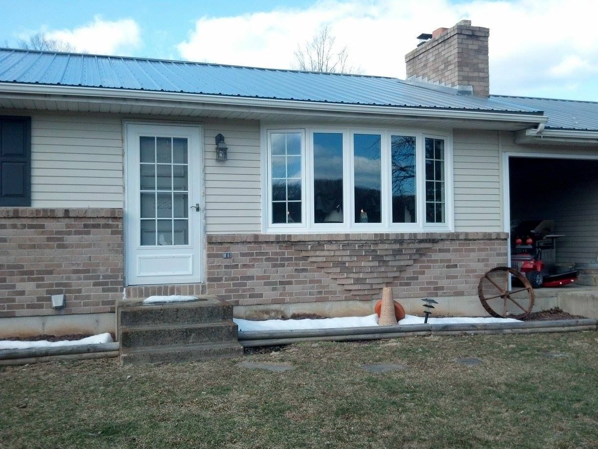 House exterior with light-colored siding, brickwork, and a metal roof. A front door and bay window are visible.