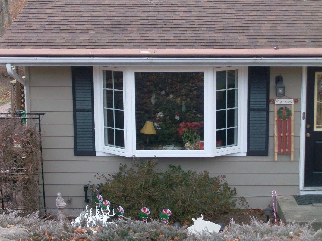 House exterior with bay window and Christmas decorations visible inside.  Tan siding, black shutters, brown roof.