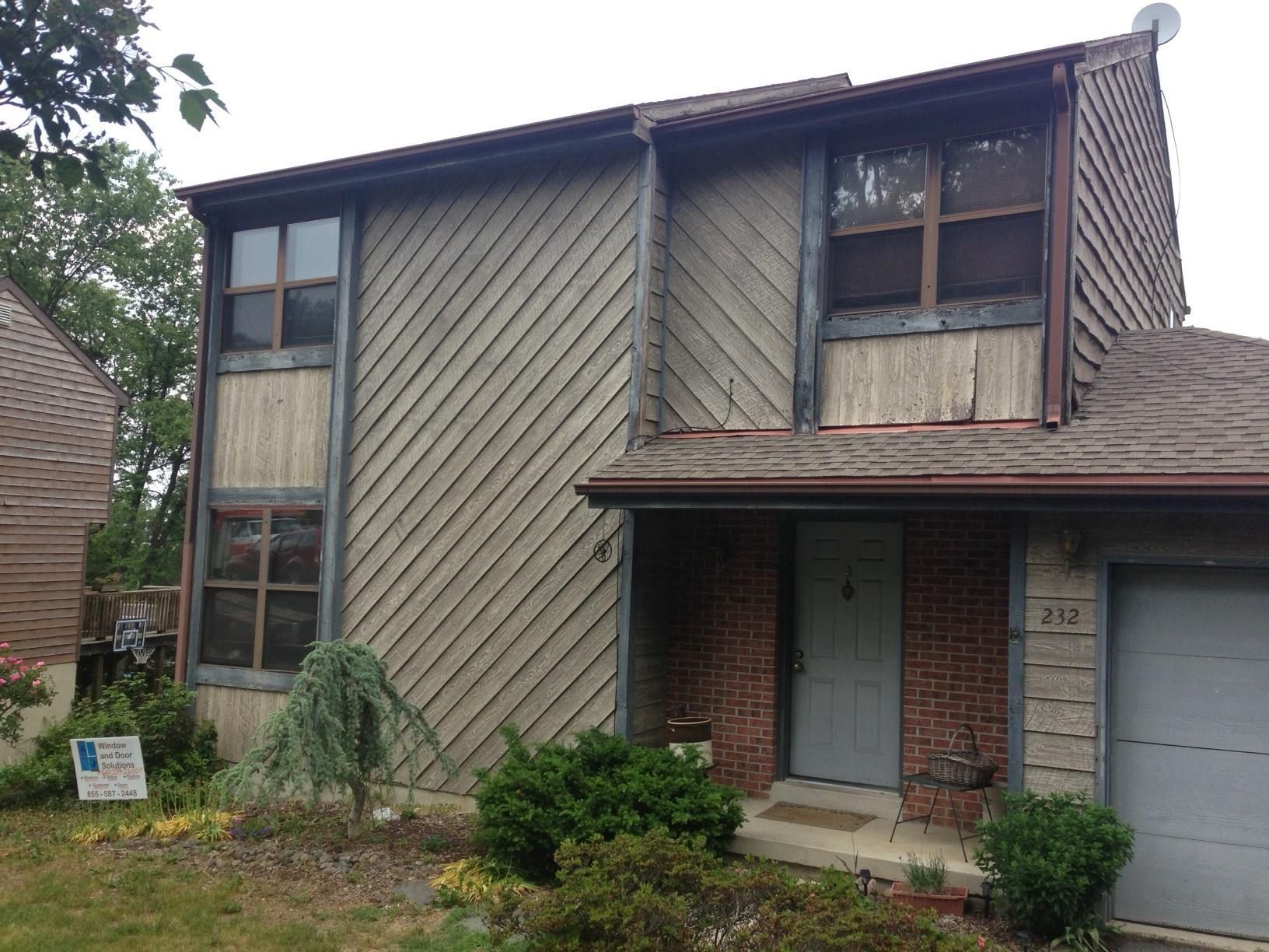 Two-story house with brown siding, brick accents, and a garage door; bushes in front.