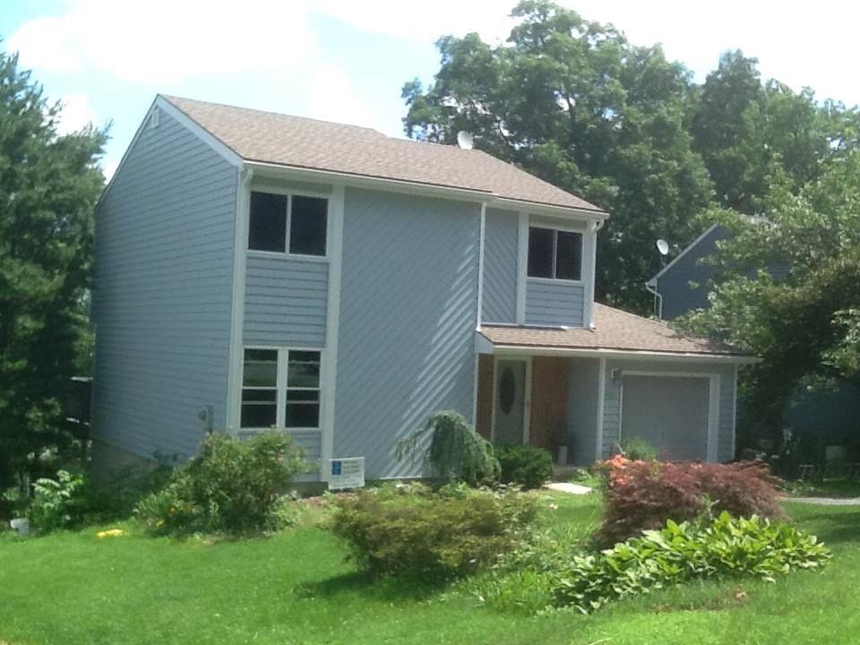 Two-story blue house with a brown roof and a small garage, surrounded by green grass and trees.